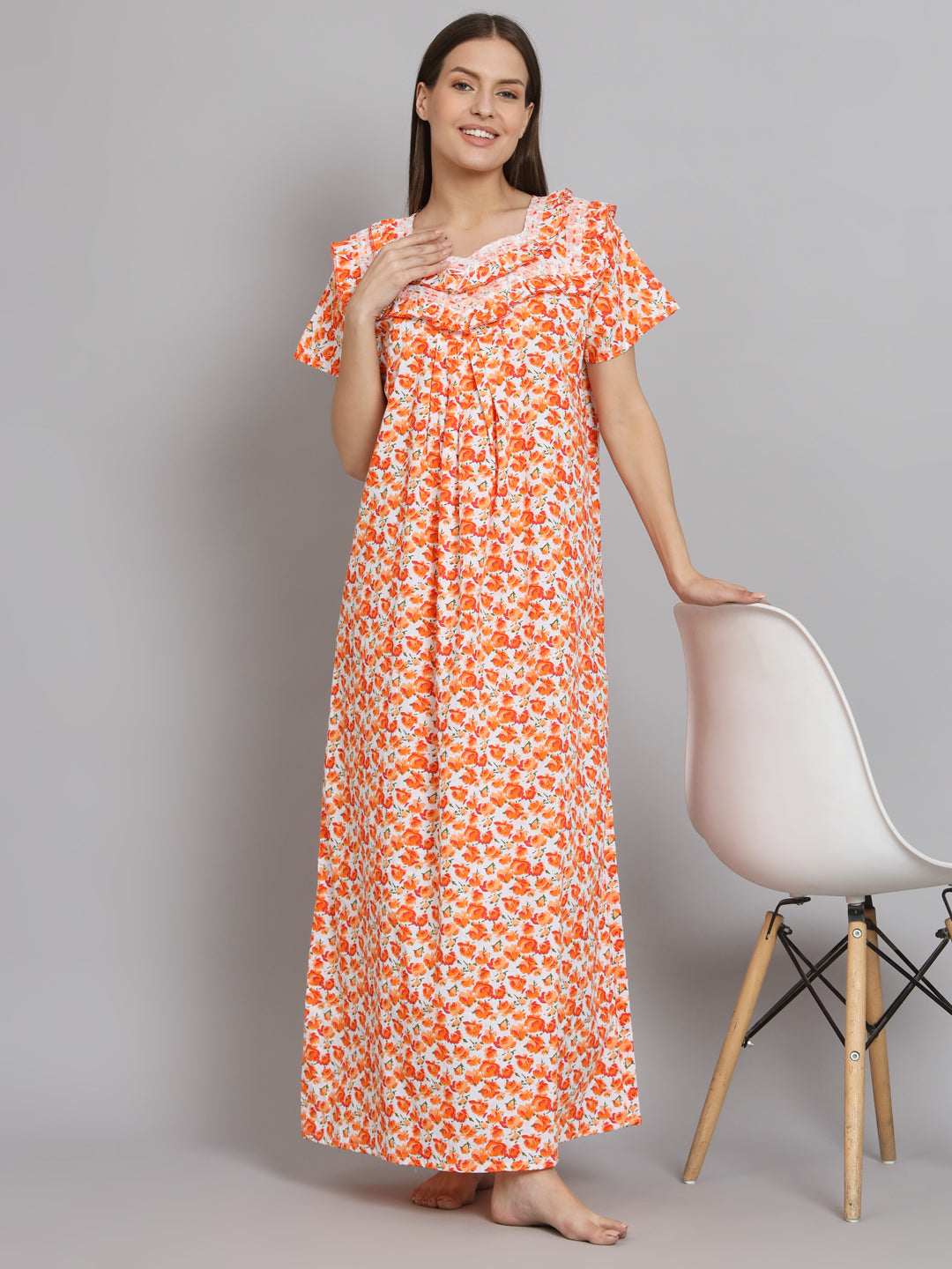 A woman wearing a long-sleeved, floor-length maxi dress with an orange and white floral pattern, standing next to a white chair against a gray studio background.