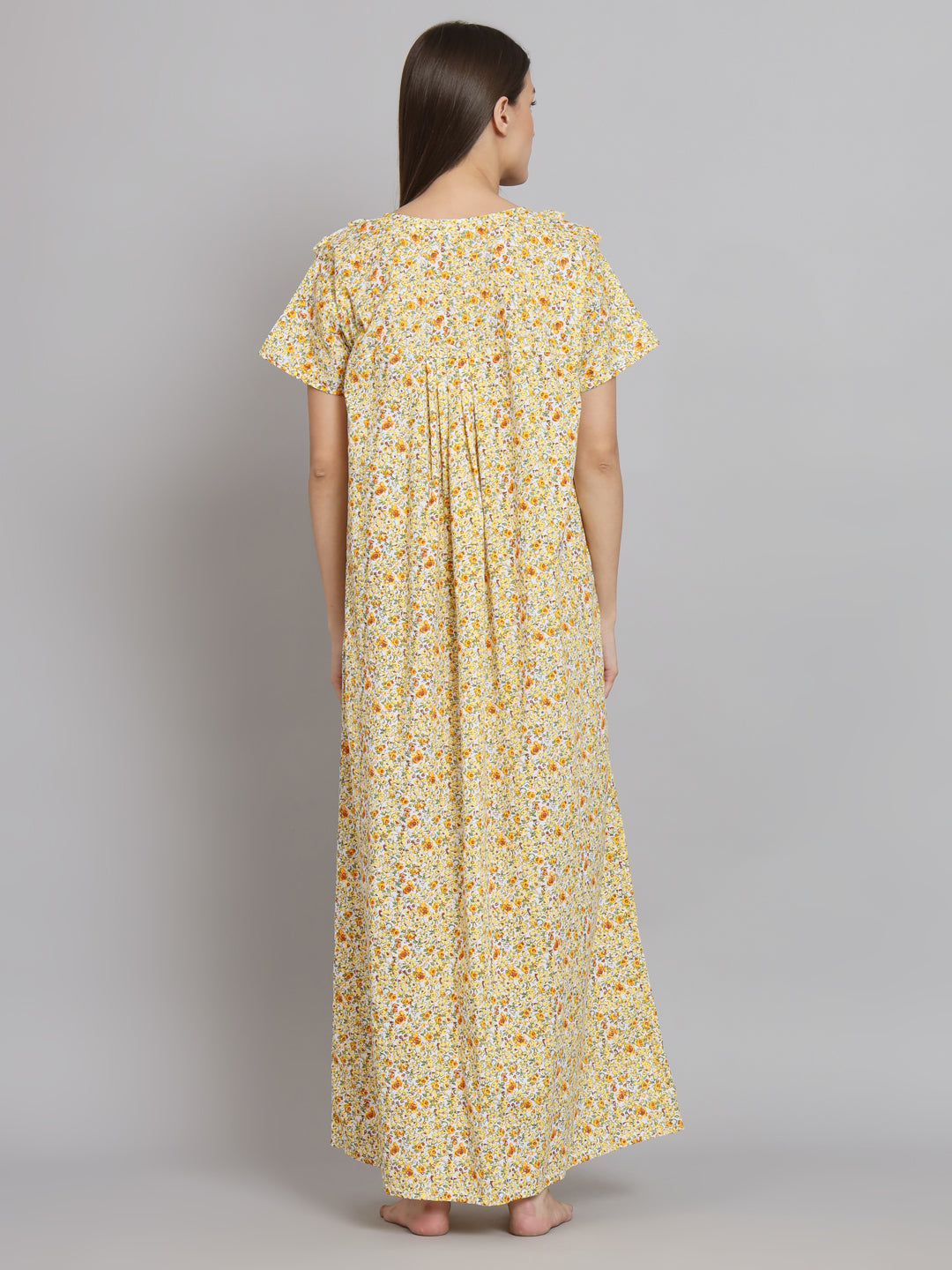 A woman viewed from the back wearing a loose, short-sleeve maxi dress with a yellow and orange floral print on a light cream background, set against a plain gray studio backdrop.