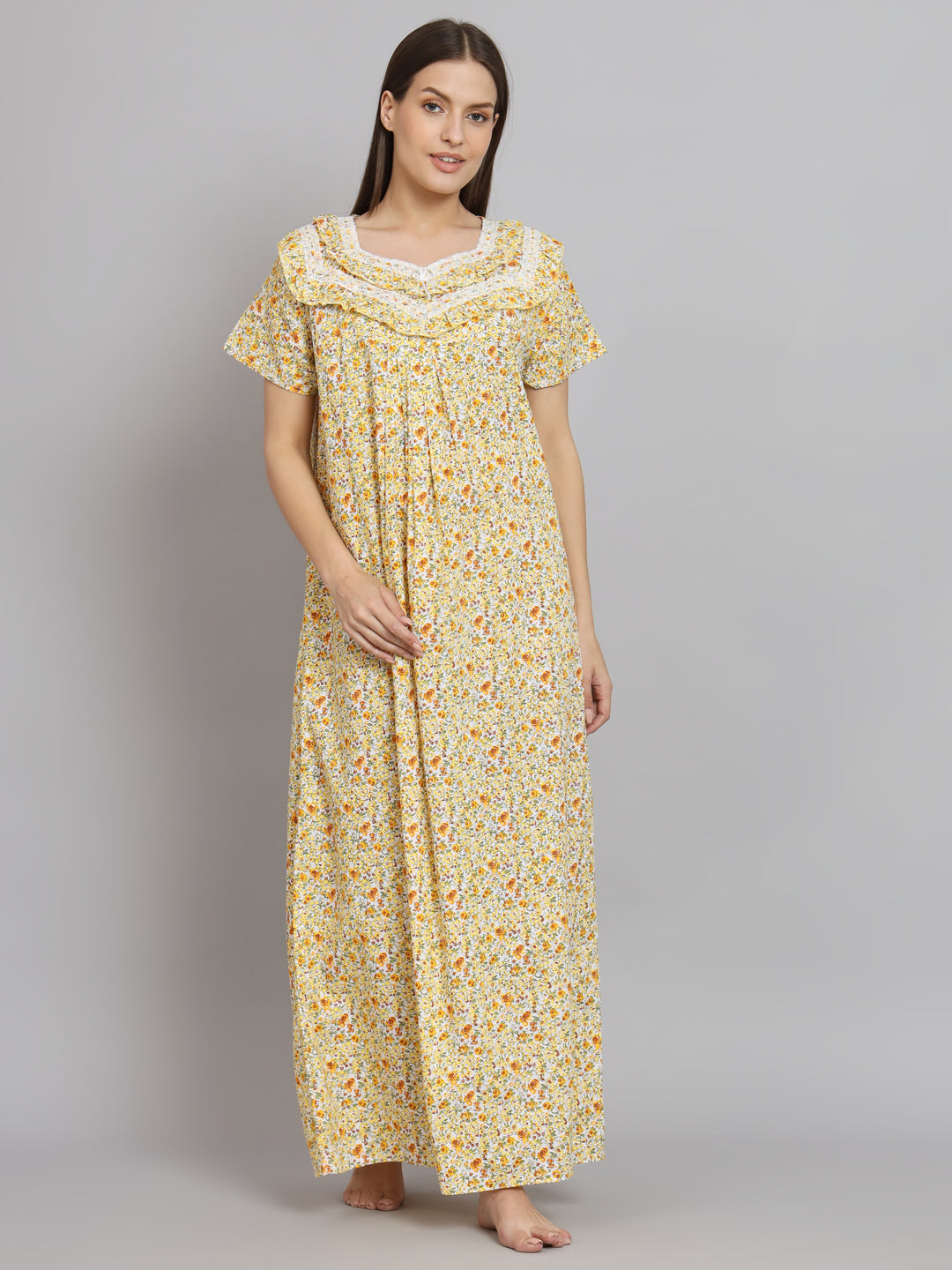 A woman modeling a long yellow nightgown with a small orange and green floral print, featuring short flutter sleeves and a lace-trimmed neckline, set against a plain gray studio background.