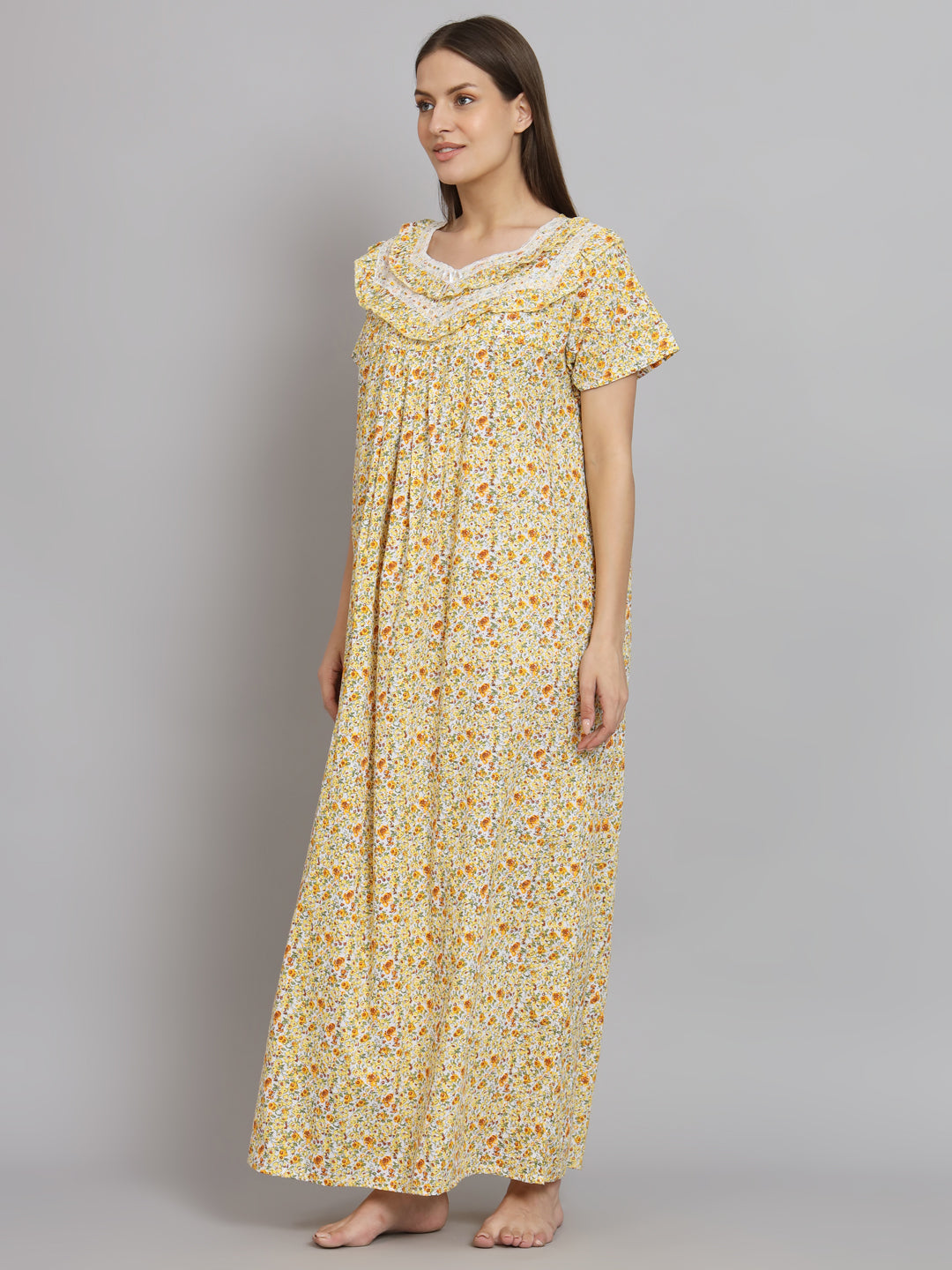 A woman modeling a long, short-sleeved nightgown with a yellow and orange floral pattern on a white base, featuring a ruffled neckline with white lace trim, set against a plain gray studio background.