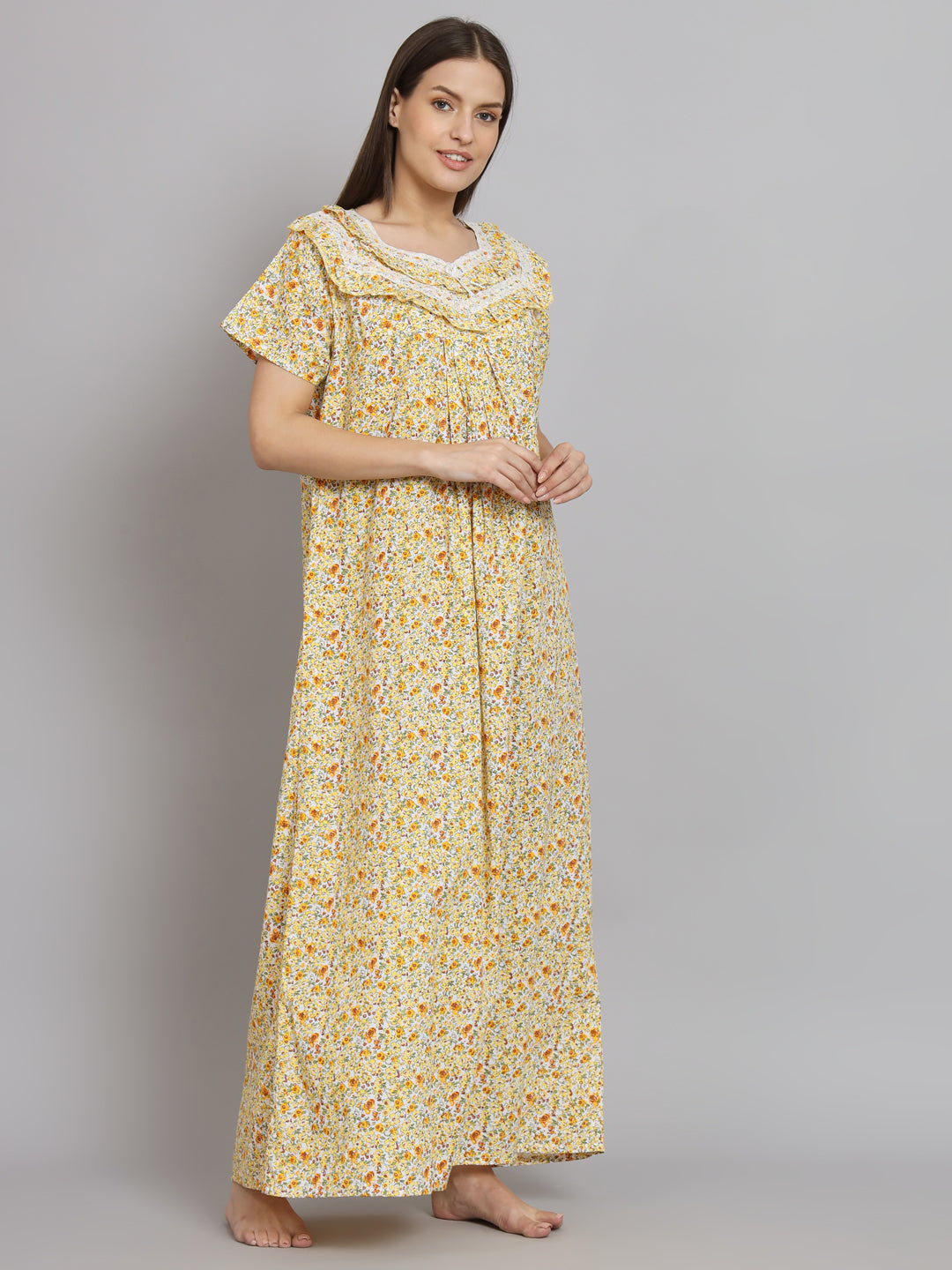 A woman modeling a long yellow floral nightgown with short sleeves and a gathered waist, standing against a plain gray studio background.