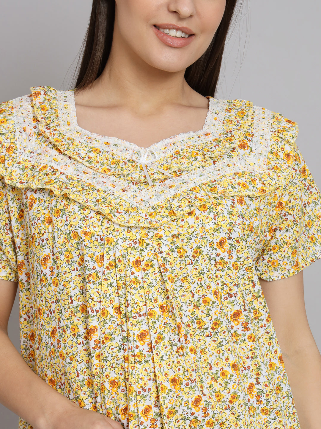 A close-up of a woman wearing a yellow and orange floral print top with a ruffled collar and white lace trim, set against a plain gray background.