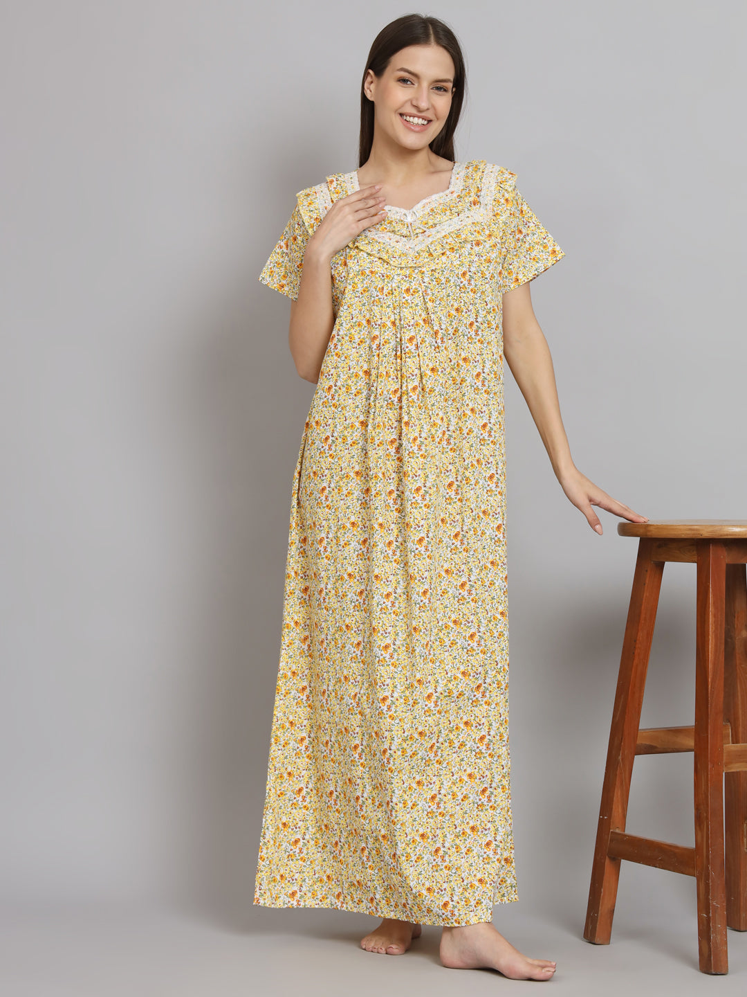 A woman modeling a yellow and orange floral print maxi nightgown with white lace trim, standing next to a wooden stool against a gray studio background.