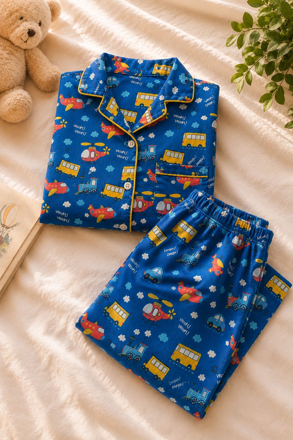 A children's two-piece pajama set laid flat on a cream fabric surface, featuring a blue long-sleeve top and matching pants with a colorful school-travel print including buses, airplanes, and helicopters. A teddy bear, book, and plant are placed around it as styling props.