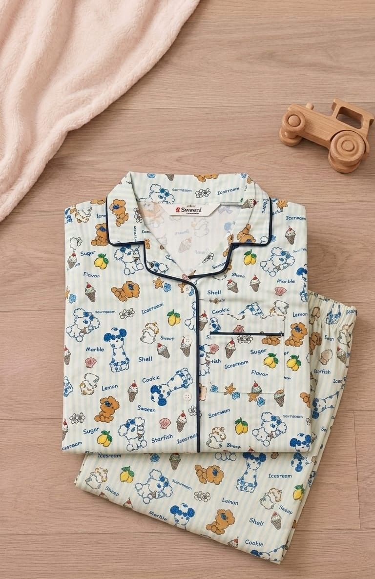 A folded set of pajama shirts and matching shorts with a cartoon dog print, styled on a light wood surface with a soft pink blanket and a small wooden toy car.