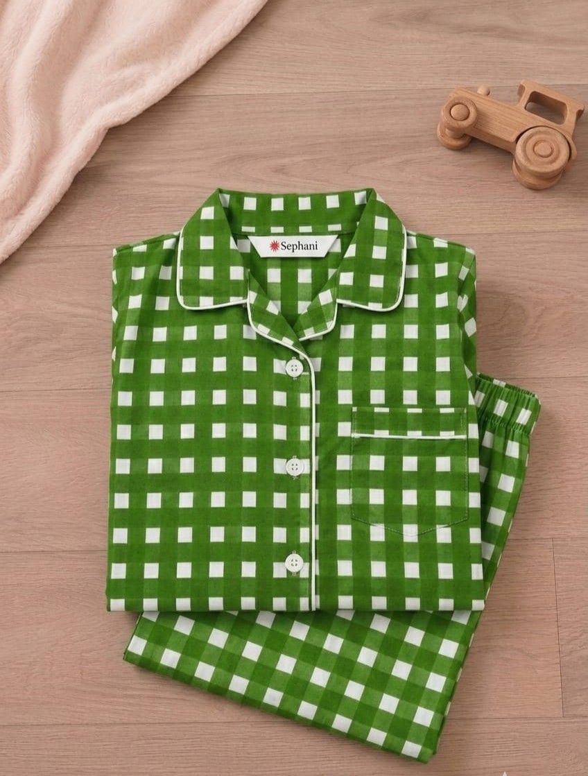 A folded green and white gingham check pajama set with a collar and button placket, placed on a light wood surface with a soft pink blanket and a small wooden toy car nearby. The brand name 'Sophani' is visible on the inner neck label.