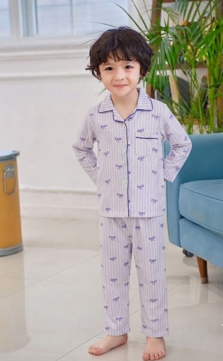 A young child wearing a light purple and white striped pajama set with small bow prints, standing in a brightly lit room with a blue sofa and green plants in the background.