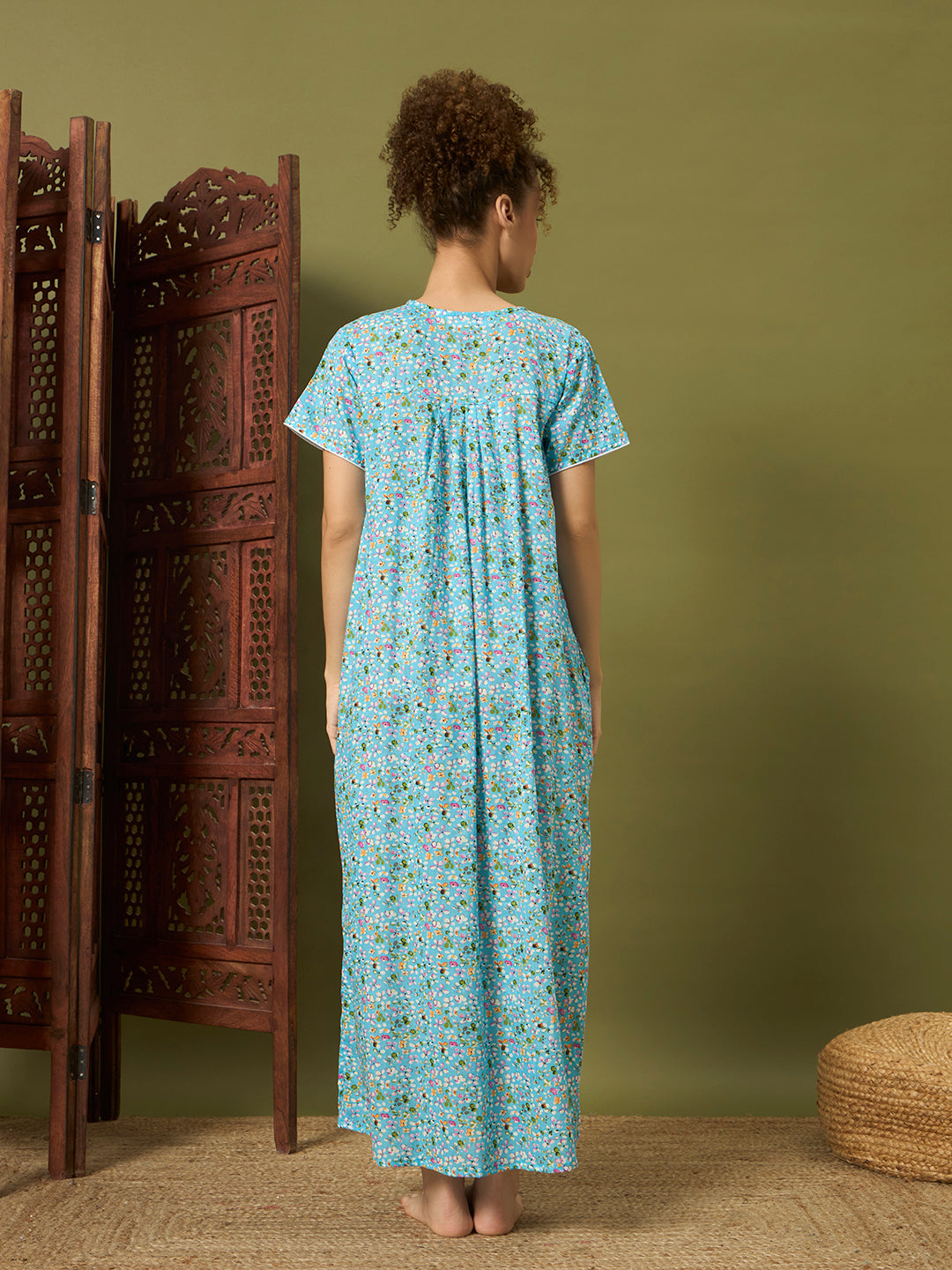 A woman stands with her back to the camera wearing a light blue short-sleeve maxi dress with a small multicolor floral print. She is standing in a styled room with a carved wooden folding screen on the left, a textured olive green wall behind her, and a woven pouf on the floor to the right.