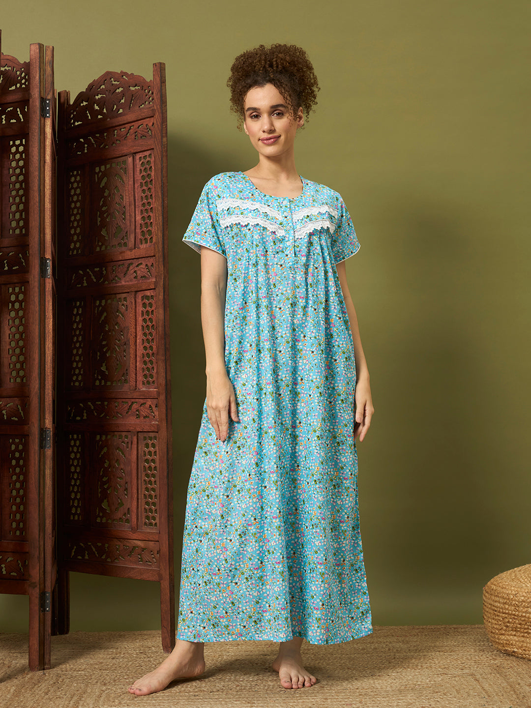 A woman stands barefoot in a room wearing a long light blue nightgown with a small floral print and white lace trim at the neckline. A carved wooden folding screen is to her left, and a muted olive-green wall is behind her.