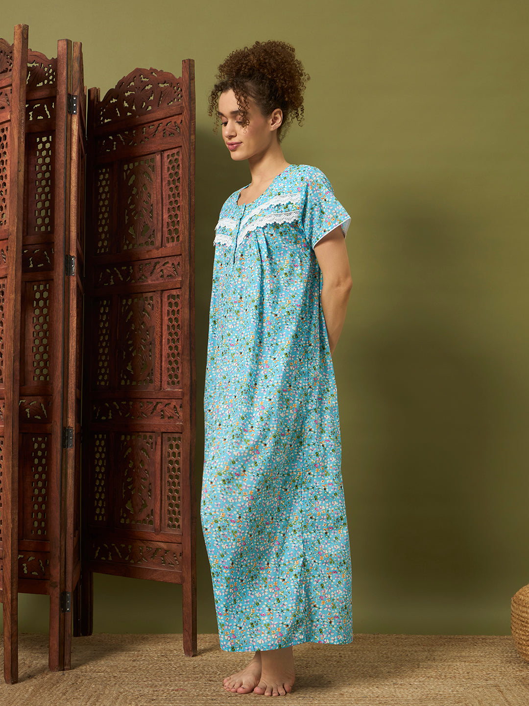 A woman stands barefoot indoors wearing a loose, short-sleeve nightgown with a light blue base and small multicolor floral print. She is posed beside a carved wooden folding screen against an olive green wall.