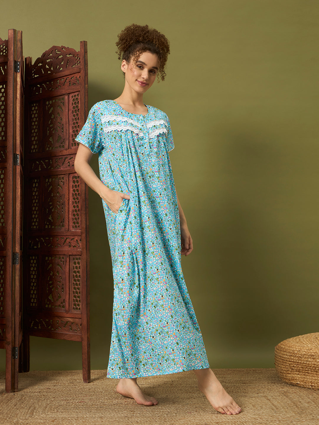 A woman stands barefoot in a bedroom-like setting wearing a light blue floral nightgown with white lace trim at the neckline. A carved wooden screen is to her left, and a woven pouf is partially visible at the lower right.