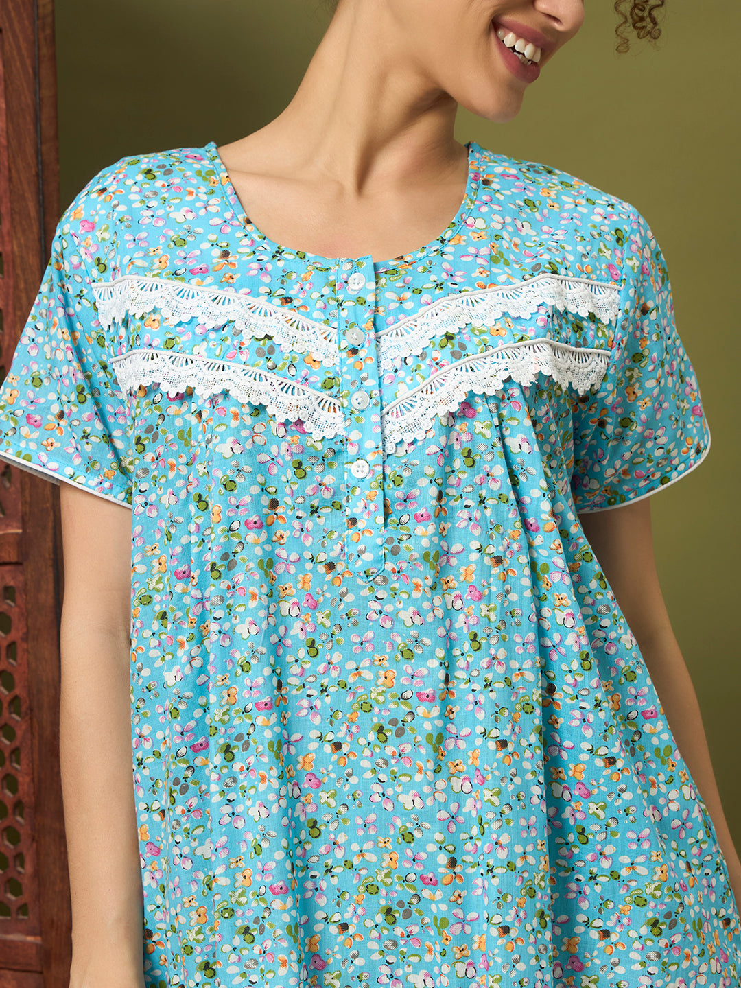 A woman wearing a light blue short-sleeve nightgown with a dense multicolor floral print and white lace trim across the chest.