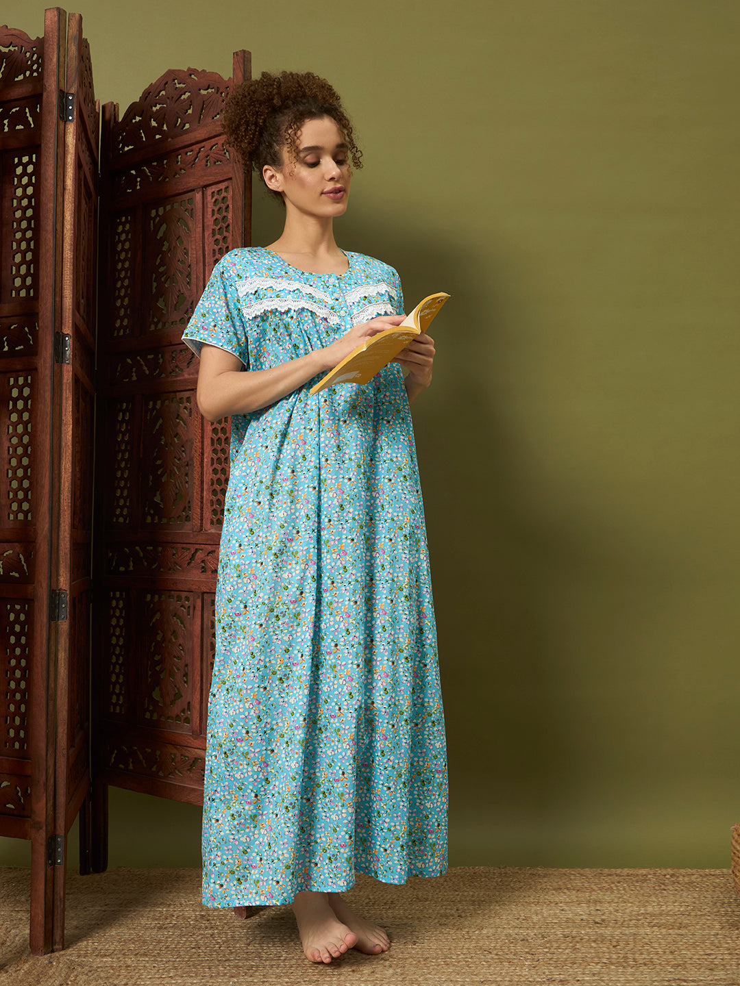 A woman stands barefoot in a studio setting, holding an open book and wearing a light blue floral nightgown with white lace trim at the neckline. She is positioned in front of a dark brown carved folding screen and a muted olive-green background.