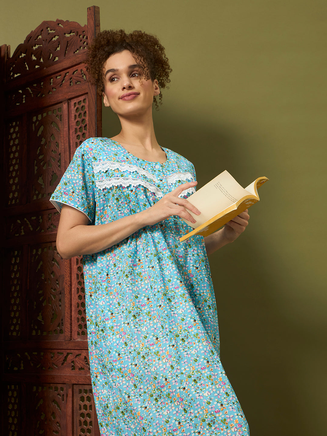 A woman with curly brown hair wearing a light blue floral nightgown stands next to a carved wooden screen, holding an open book against an olive green background.