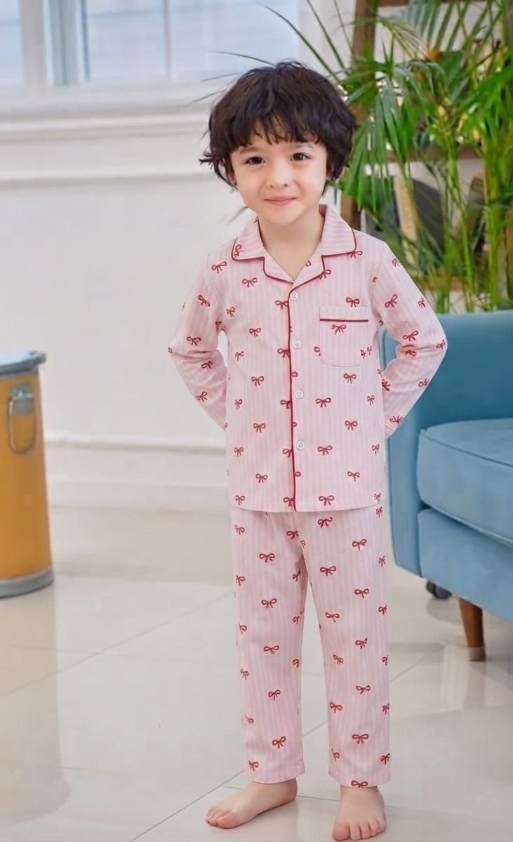 A young child wearing a pink long-sleeve pajama set with a red bow pattern, standing barefoot in a bright indoor room with a blue sofa and green plant in the background.