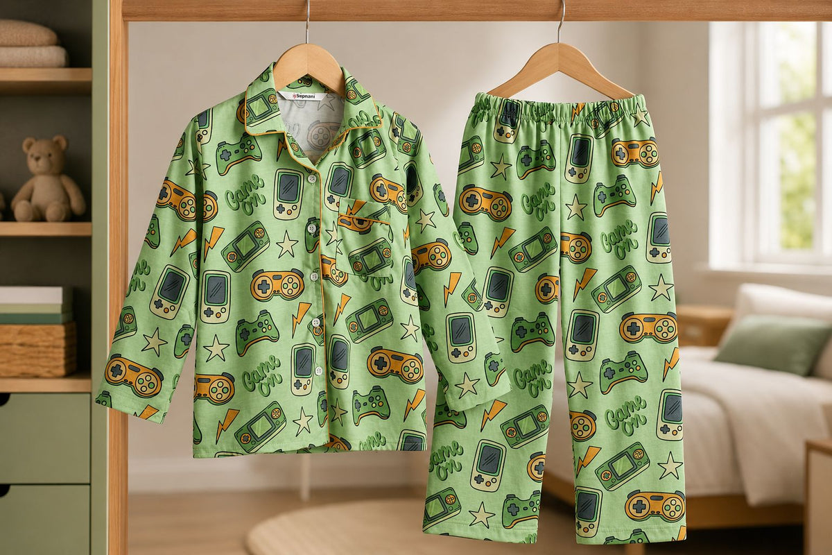 A two-piece kids' pajama set hanging on wooden hangers in a bedroom setting. The set features a light green long-sleeve button-up shirt and matching pants, both printed with a colorful retro video game controller pattern.