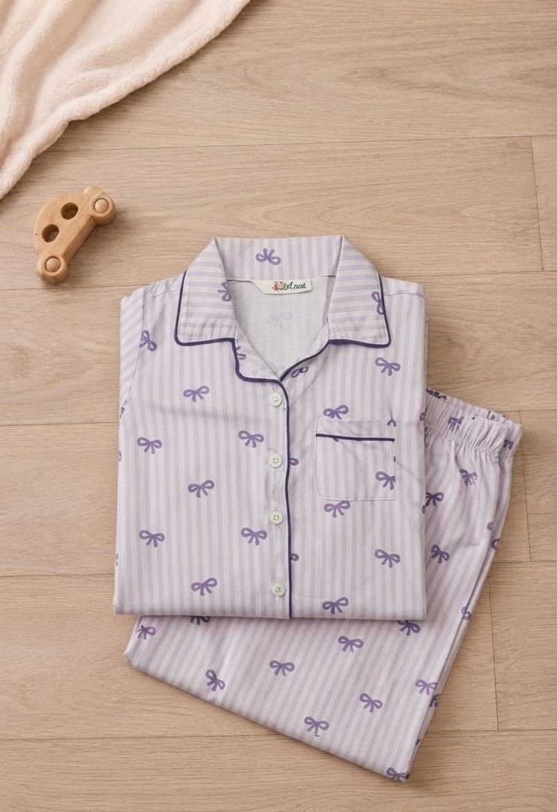 A folded pajama set consisting of a long-sleeve button-up shirt and matching pants, laid flat on a light wood floor. The pajamas are light lavender with thin white vertical stripes, purple bow motifs, and dark purple piping.