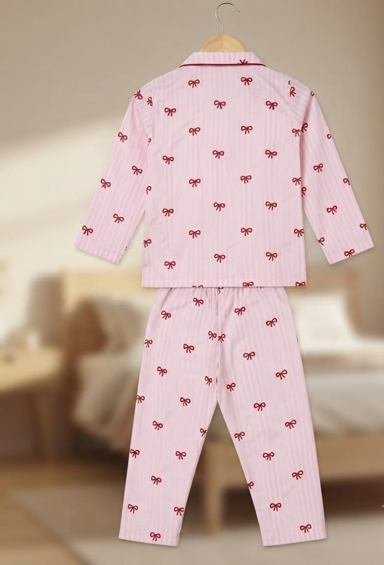 A two-piece children's pajama set consisting of a long-sleeve top and matching pants, shown hanging against a softly blurred bedroom background. The pajamas are light pink with thin vertical stripes and a repeating pattern of small red bows.
