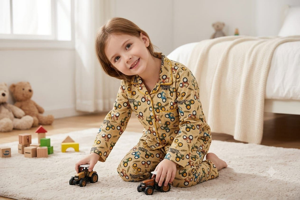 A young girl with light brown hair sits on a cream rug in a brightly lit bedroom, wearing a yellow long-sleeve pajama set with a colorful pattern of cartoon vehicles. She is holding two toy trucks.