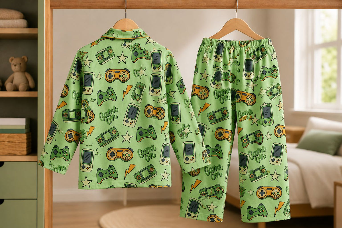 A two-piece pajama set hanging on wooden hangers, featuring a light green long-sleeve top and matching pants with a retro handheld game console print. The print includes dark green game controllers, orange star rockets, and white stars with 'Game On!' text.