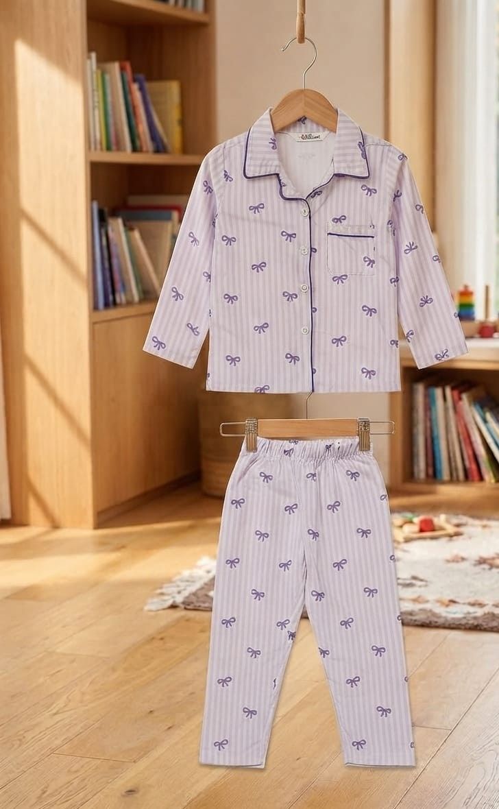 A two-piece children's pajama set hanging on wooden hangers, featuring a long-sleeve top and matching pants in a light lavender-white color with thin vertical stripes and a repeating bow print. The set is displayed in a warm, sunlit child's bedroom with wooden floors and bookshelves in the background.
