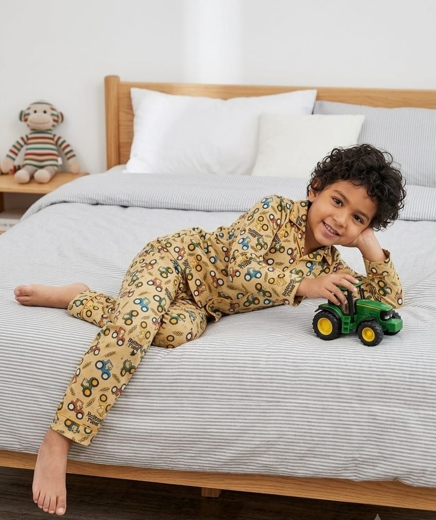 A young boy with curly dark hair lies on a bed wearing a beige long-sleeve pajama set with a colorful tractor and bicycle pattern. He is holding a green toy tractor, with a wooden headboard, white pillows, and a striped sheet in the background.