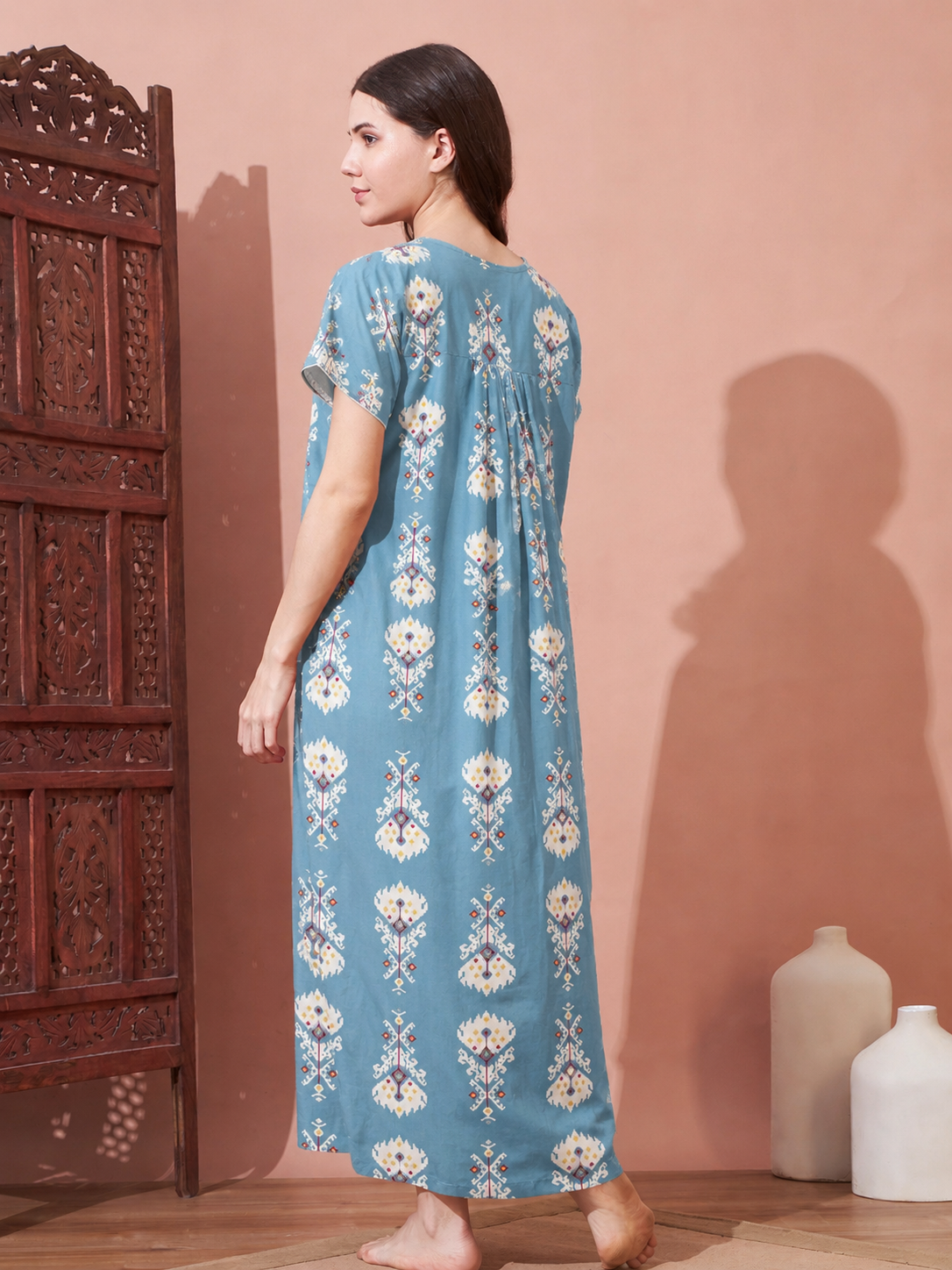 A woman stands barefoot in profile wearing a long, loose-fitting blue nightgown with cream and white floral embroidery, set against a warm pinkish-brown wall with a wooden screen and white ceramic vases.