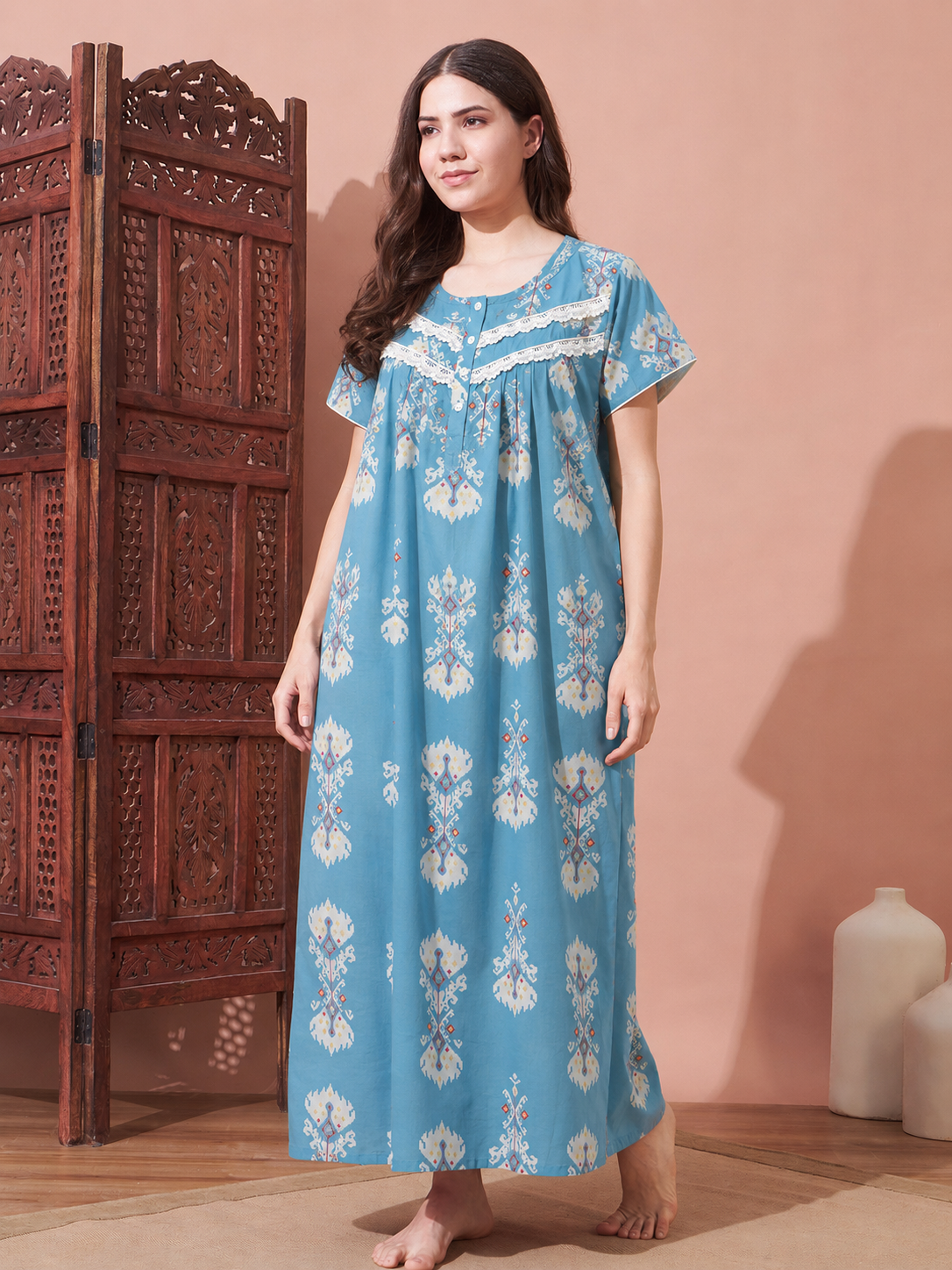 A woman stands barefoot in a bedroom-like setting wearing a long light blue nightgown with white floral prints and lace trim at the neckline. A carved wooden screen is to her left, and two white ceramic bottles sit on the floor to her right against a peach-pink wall.