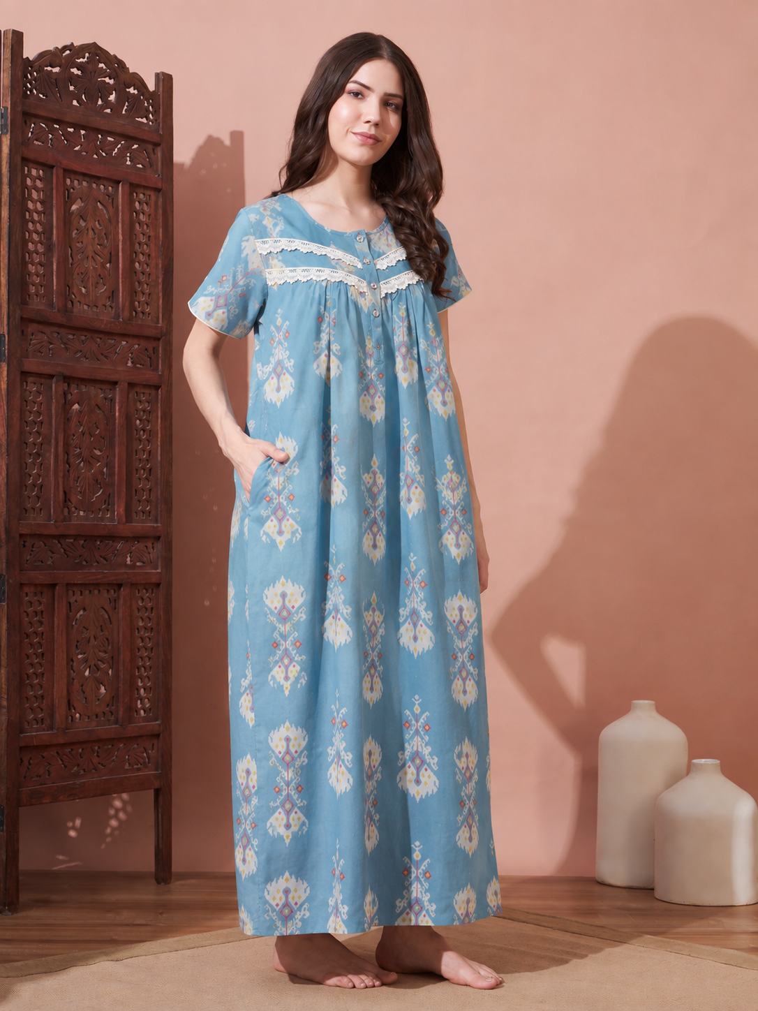 A woman stands barefoot in a room wearing a light blue cotton nightgown with white and beige floral patterns, lace trim at the neckline, and side pockets. She is posed next to a carved wooden cabinet against a dusty pink wall.