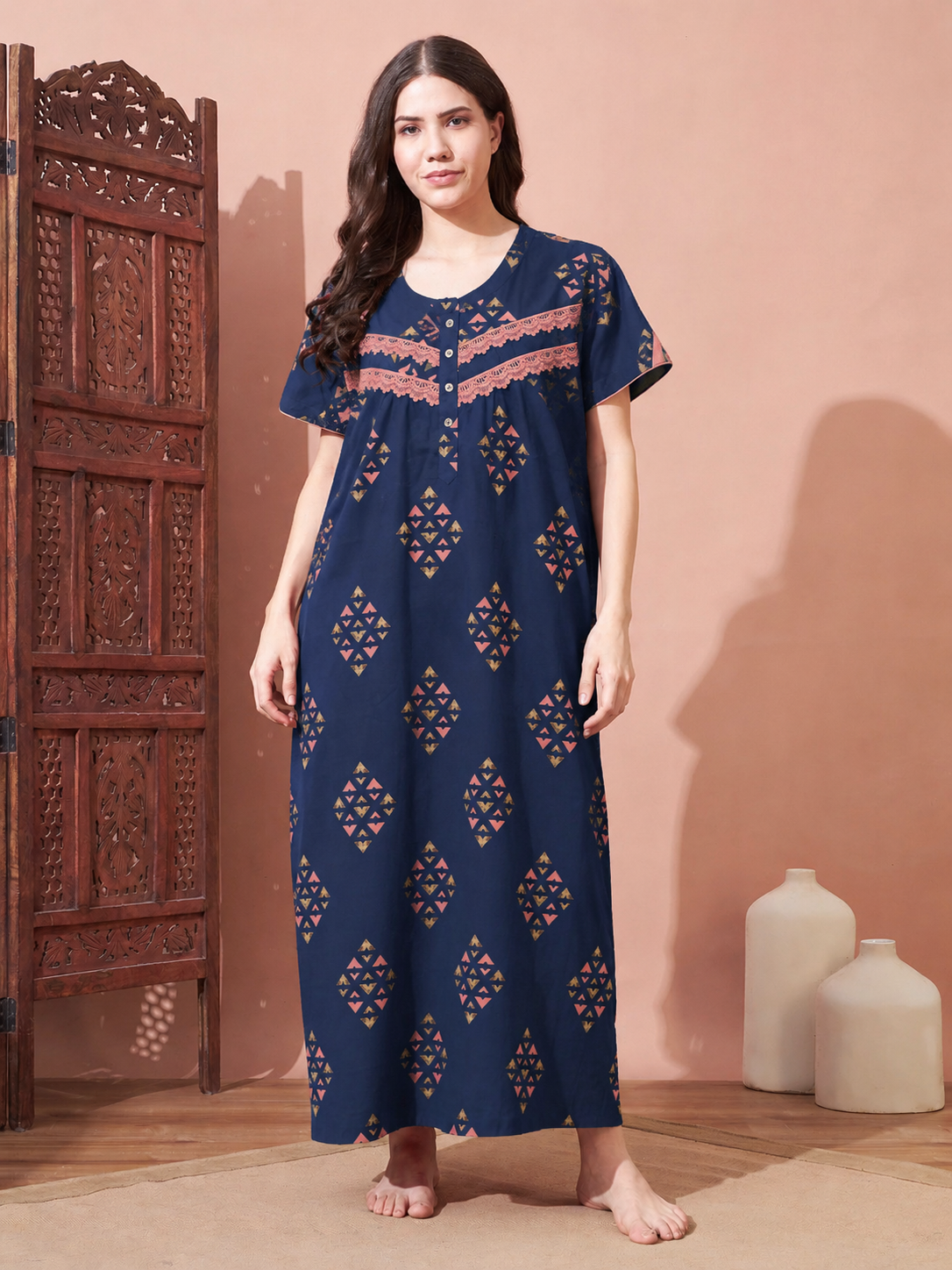A woman stands barefoot in a room wearing a navy blue maxi nightgown with orange and gold geometric patterns and a decorative neckline. She is positioned next to a carved wooden screen, with two white ceramic vases on the floor against a warm peach-colored wall.