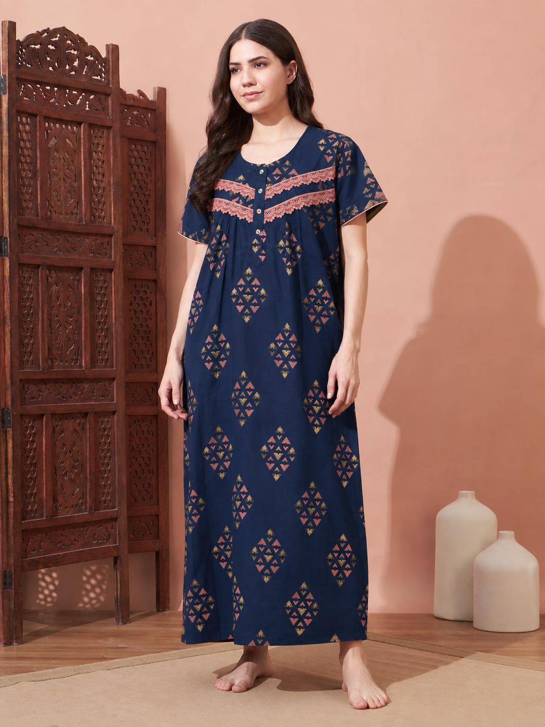 A woman stands barefoot in a studio setting wearing a navy maxi nightgown with geometric cutout patterns and a pink embroidered yoke. She is posed next to a carved wooden screen with two white ceramic vases against a peach-colored wall.