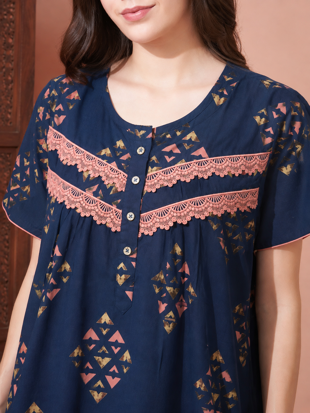A woman wearing a navy blue short-sleeve tunic top with pink lace trim and geometric gold patterns, shown from the chest up against a warm peach-colored background.
