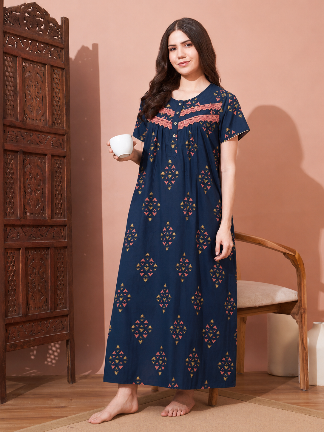 A woman stands barefoot in a room wearing a navy blue maxi nightgown with orange and yellow geometric patterns and a pink embroidered neckline. She holds a small white cup next to a carved wooden cabinet and a wooden chair.