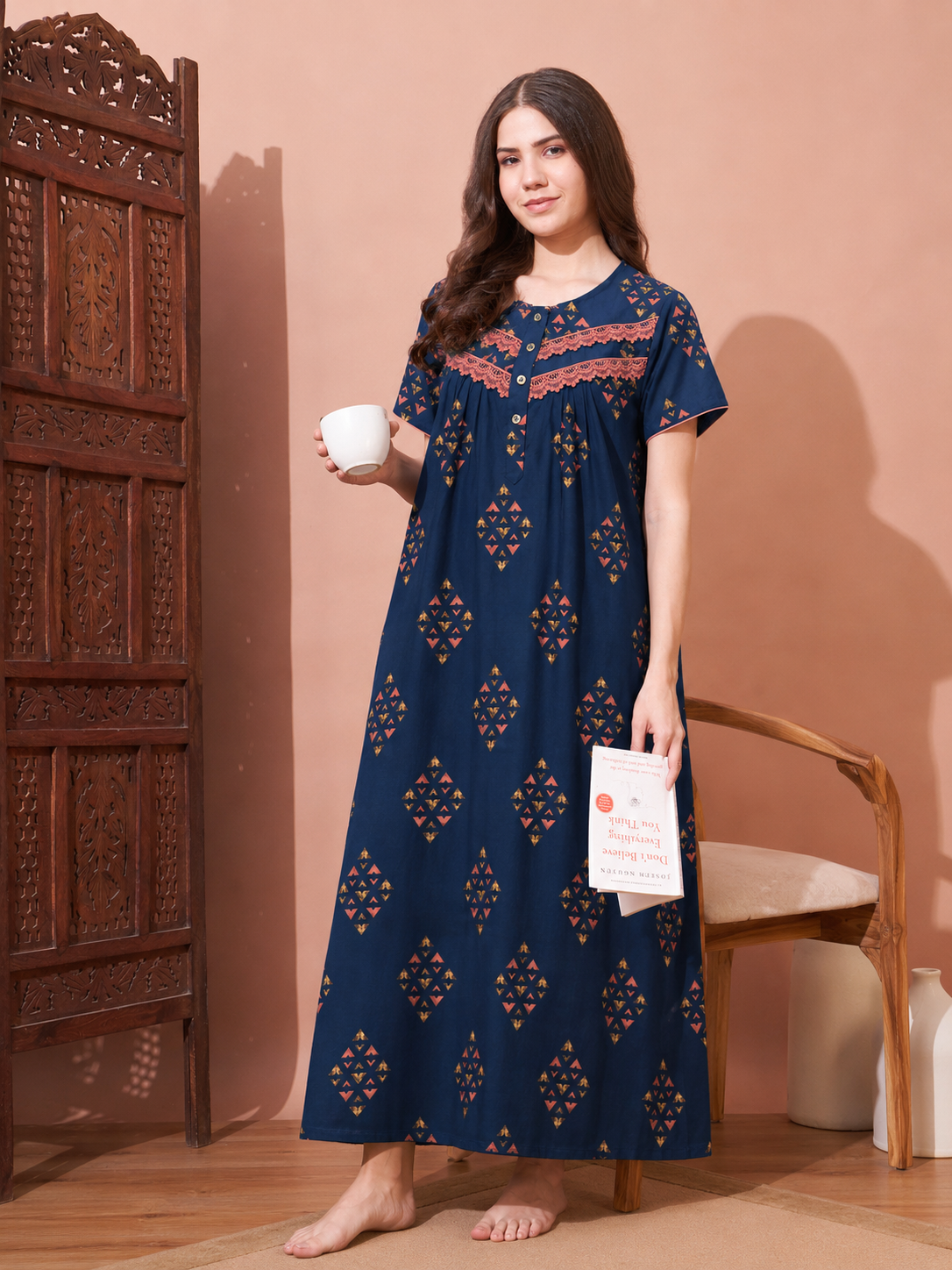 A woman stands barefoot in a room wearing a navy blue nightgown with orange and gold geometric patterns and embroidered detailing around the neckline. She holds a white mug in one hand and a book in the other, standing next to a carved wooden cabinet and a wooden chair against a warm peach-colored wall.