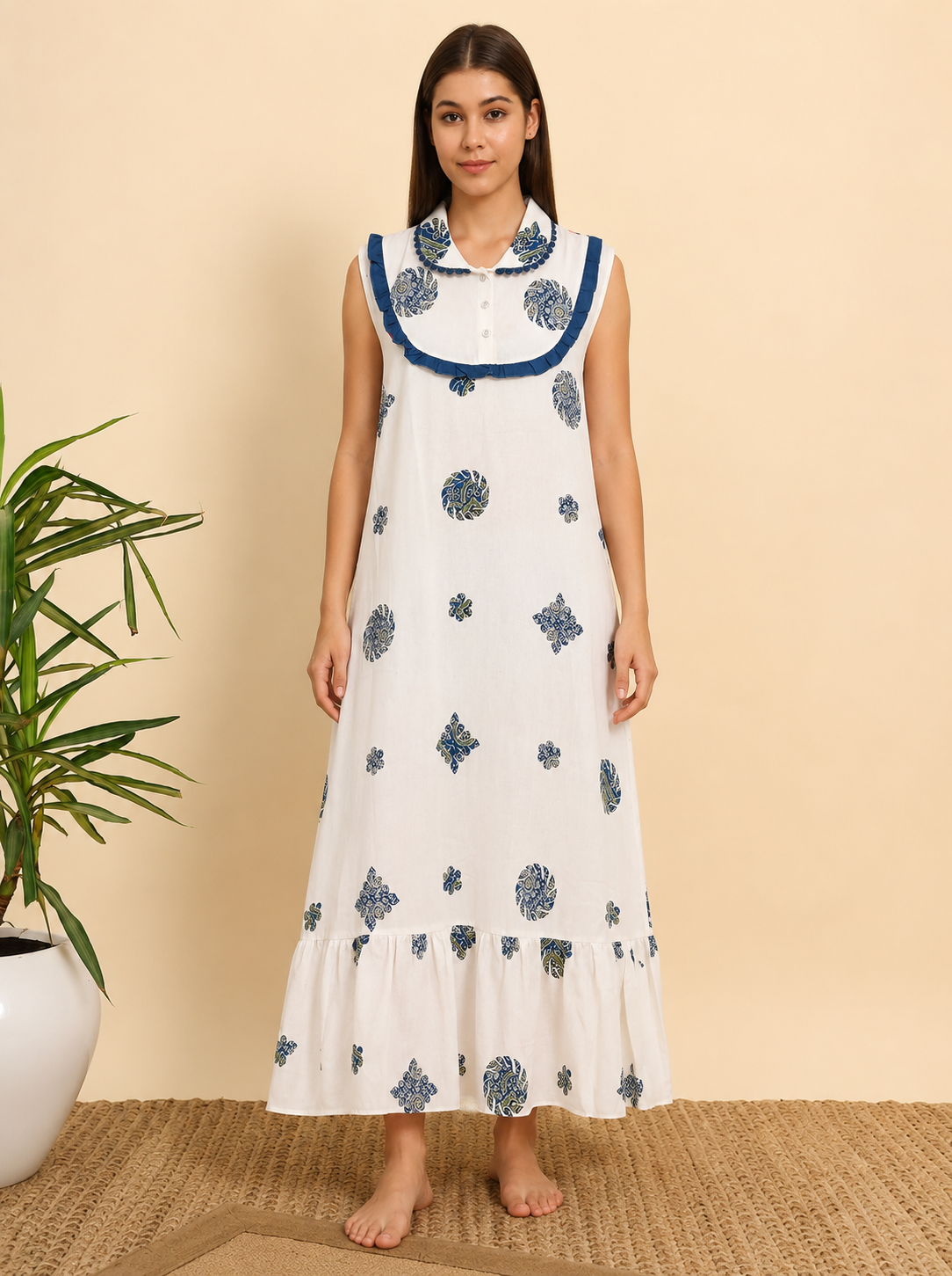 A woman stands barefoot indoors wearing a sleeveless white maxi dress with blue floral block prints and a blue collar trim. The background is a warm beige wall with a woven rug and a potted plant on the left.
