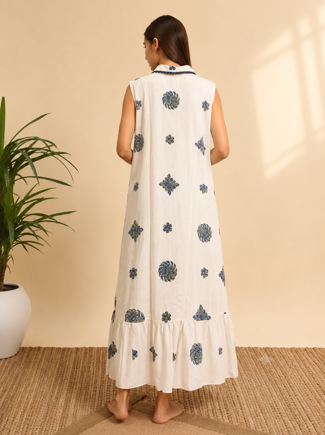 A woman stands with her back to the camera wearing a sleeveless, off-white maxi dress with blue and gray block-print style motifs. The dress features a tiered ruffle hem and a matching off-white collar with blue trim.