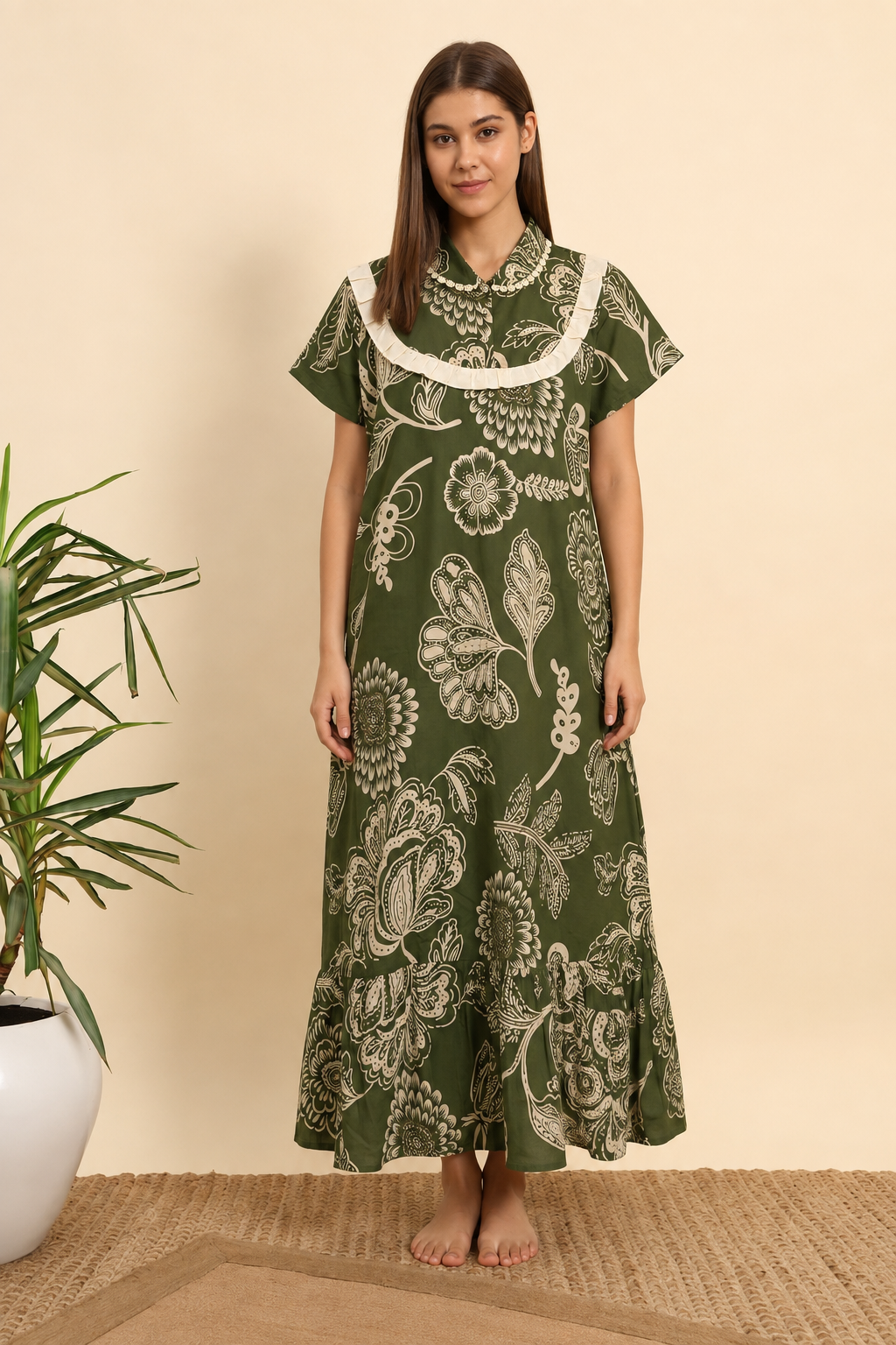 A woman stands barefoot indoors wearing a long green dress with a large white floral print and a white ruffled collar. A potted plant is visible at the left edge against a warm beige wall.