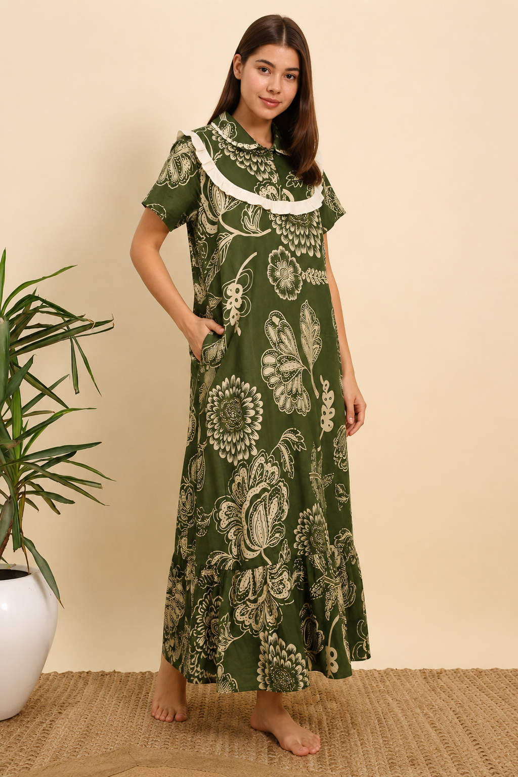A woman modeling a short-sleeved green maxi dress with a white floral and butterfly print, standing barefoot on a woven rug against a beige wall.