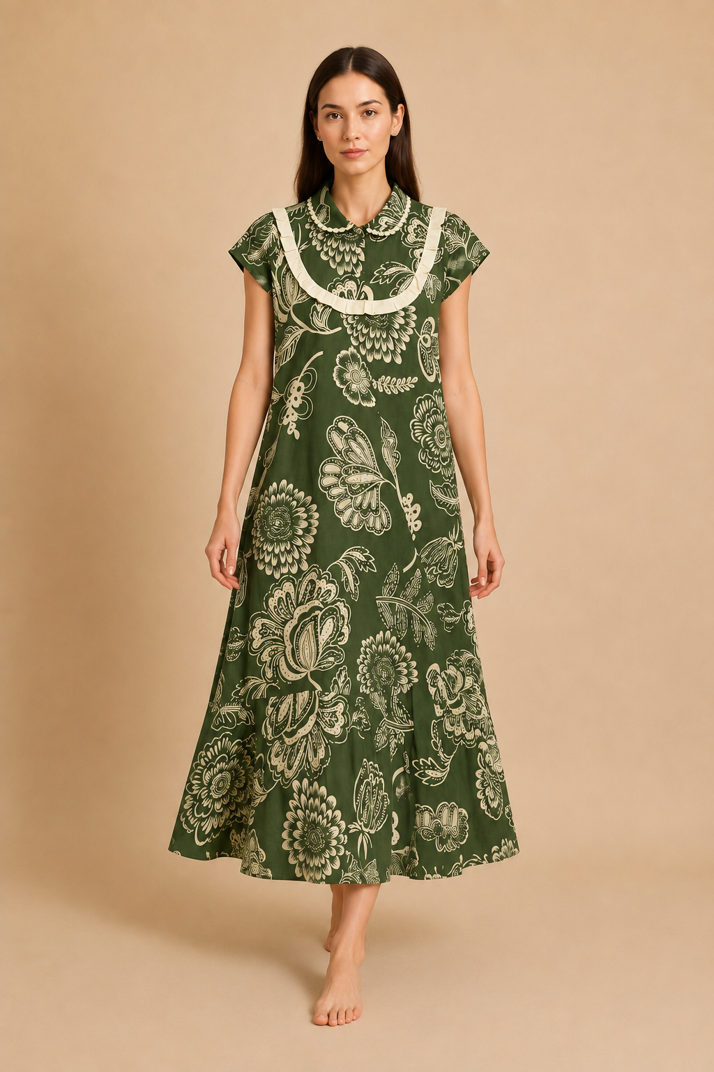 A woman stands barefoot against a plain beige studio background wearing a knee-length green dress with a large cream floral print and a cream ruffled collar.