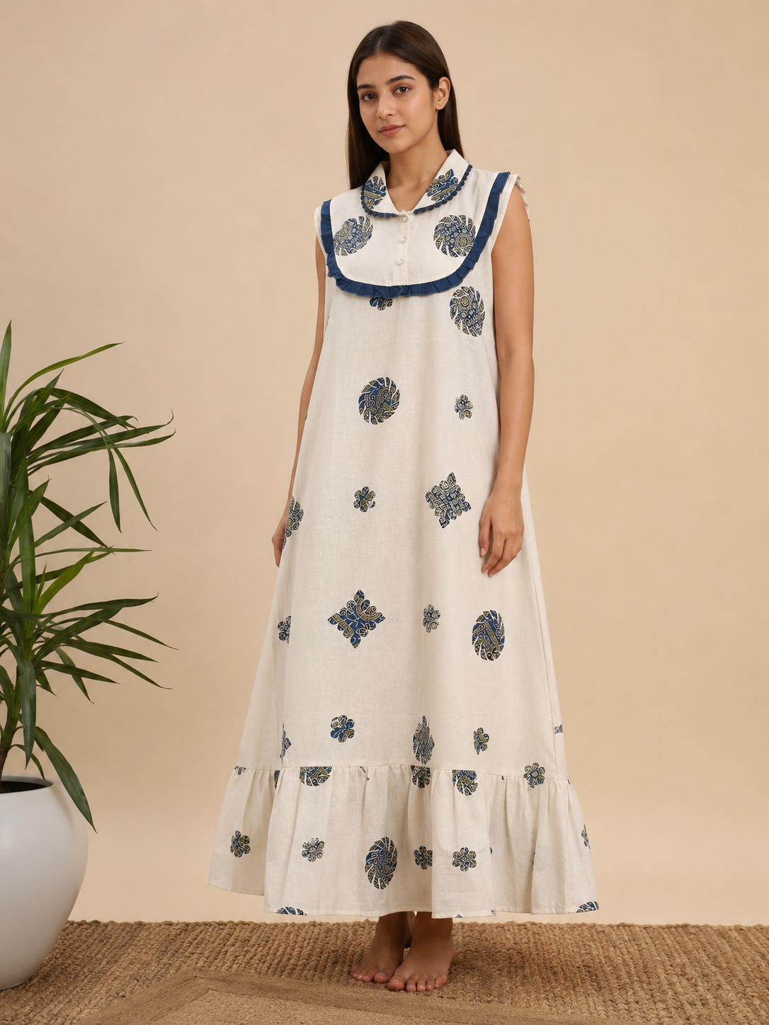 A woman stands barefoot indoors wearing a sleeveless, floor-length white dress with blue printed motifs and a blue collar trim. A potted plant and woven rug are visible against a beige wall.
