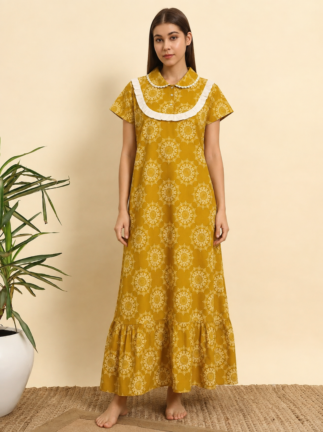 A woman stands barefoot indoors wearing a mustard yellow maxi dress with a white floral print and a white ruffled Peter Pan collar. The background is a solid beige wall with a potted plant on the left.