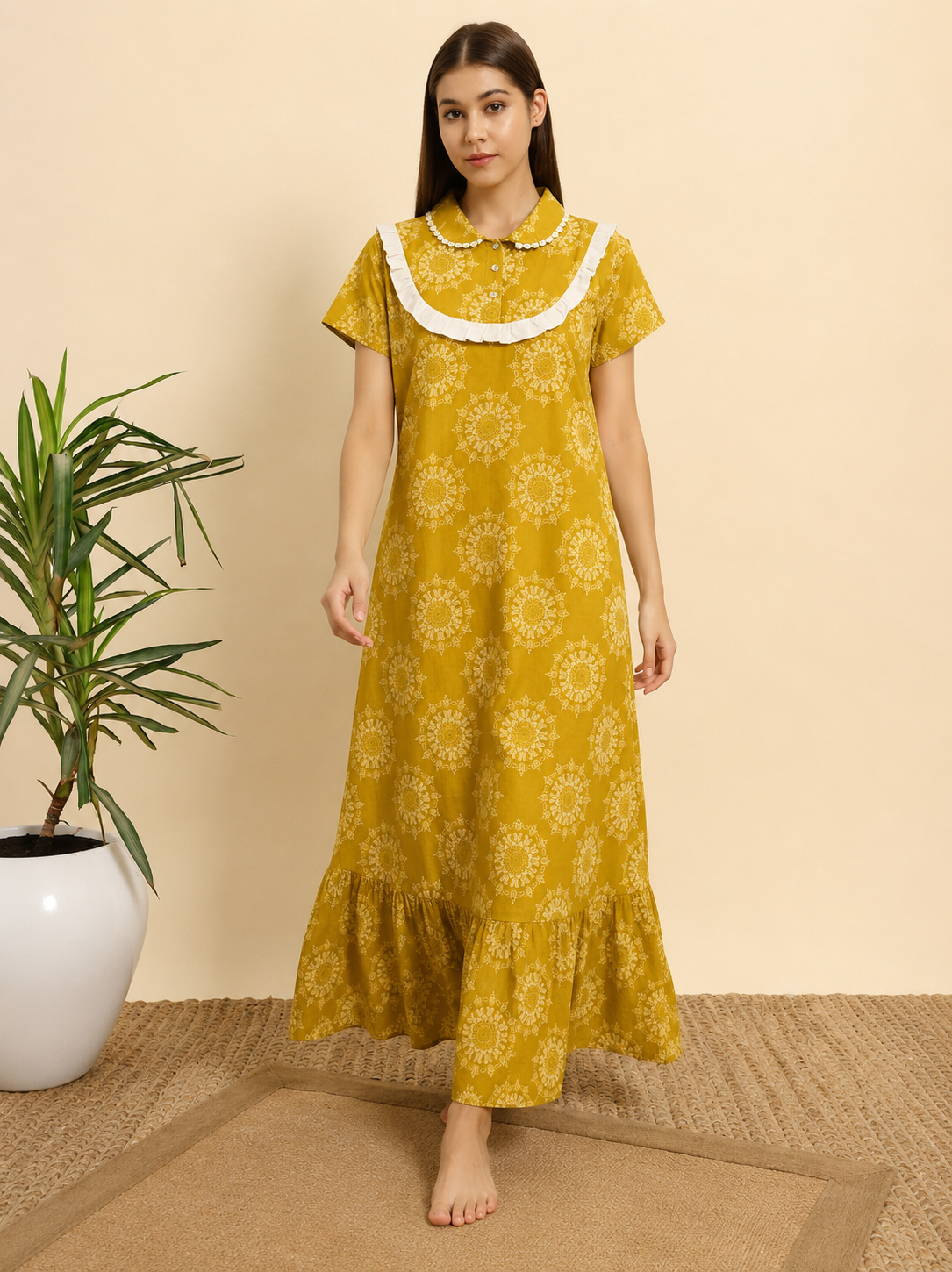 A woman stands barefoot indoors wearing a mustard yellow maxi dress with a white floral print and a white ruffled collar. The background is a warm beige wall with a potted plant on the left and a woven rug beneath her.