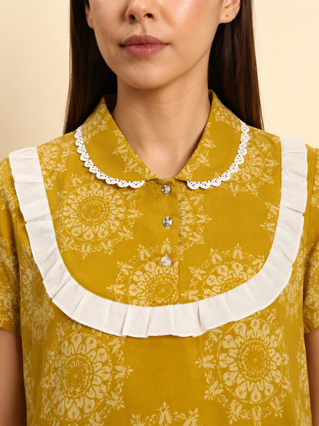 A close-up of a woman wearing a mustard yellow short-sleeve blouse with a cream floral mandala pattern, featuring a white ruffled Peter Pan collar with silver buttons.