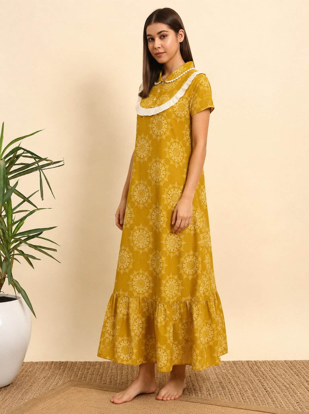 A woman modeling a mustard yellow maxi dress with a white floral mandala pattern, a white lace-trimmed high collar, and short sleeves, standing barefoot against a beige wall with a potted plant on the left.