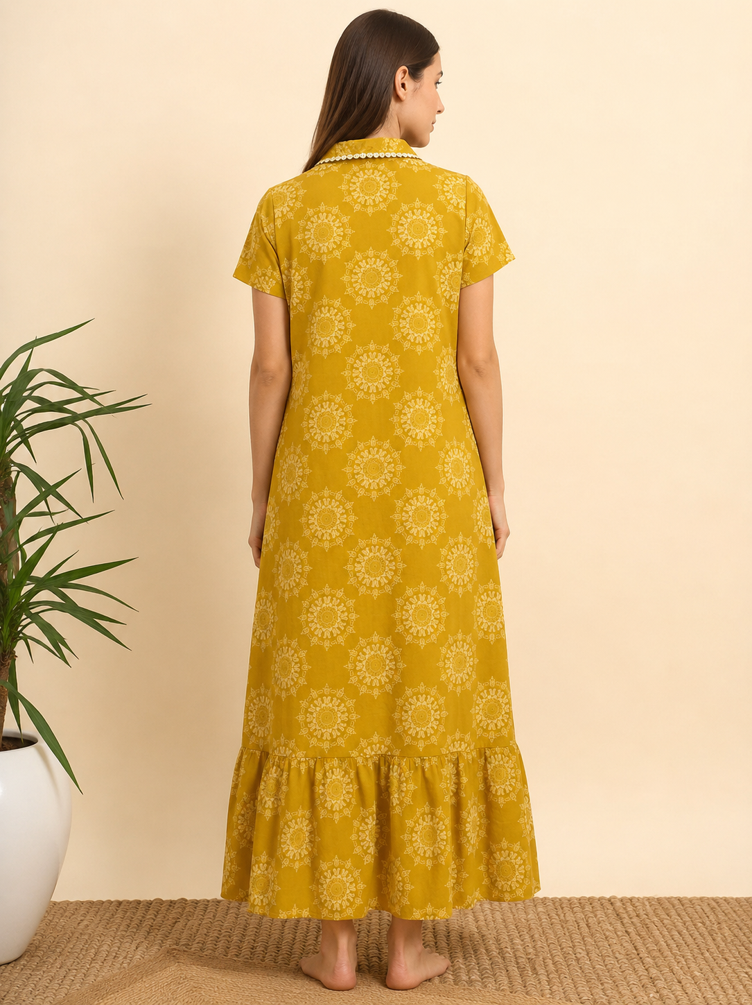 A woman viewed from the back wearing a mustard yellow short-sleeve maxi dress with a white floral mandala pattern and a ruffled hem, standing against a beige wall.