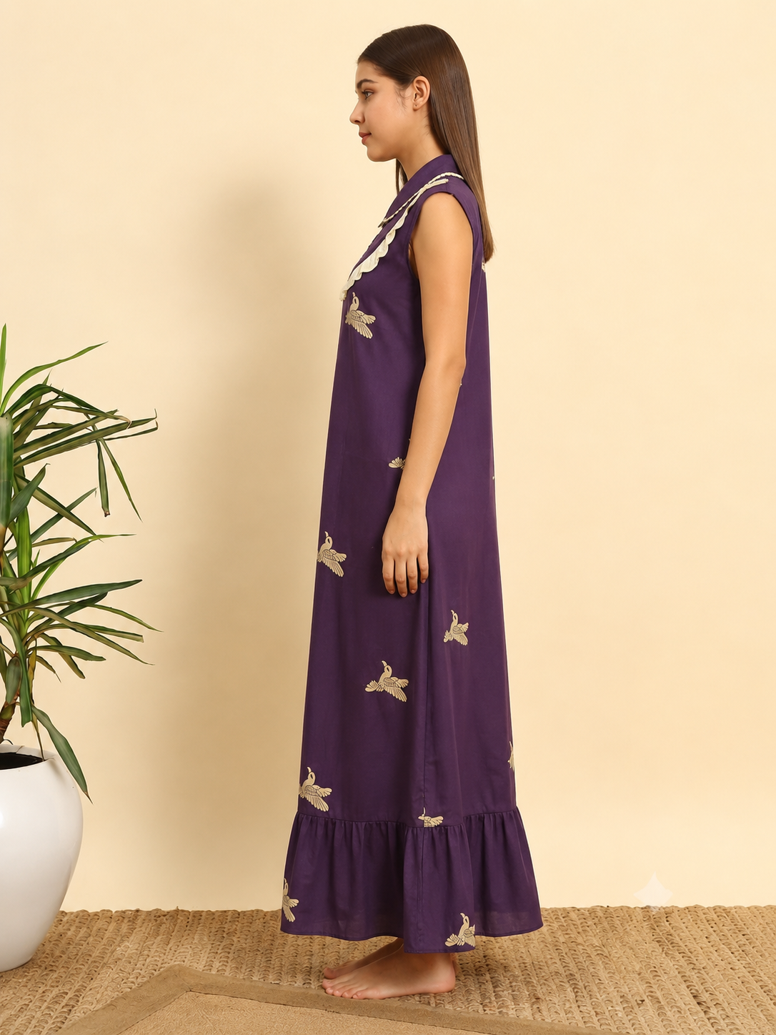 A woman wearing a sleeveless purple maxi dress with beige butterfly prints, standing in profile against a beige wall with a potted plant on the left.