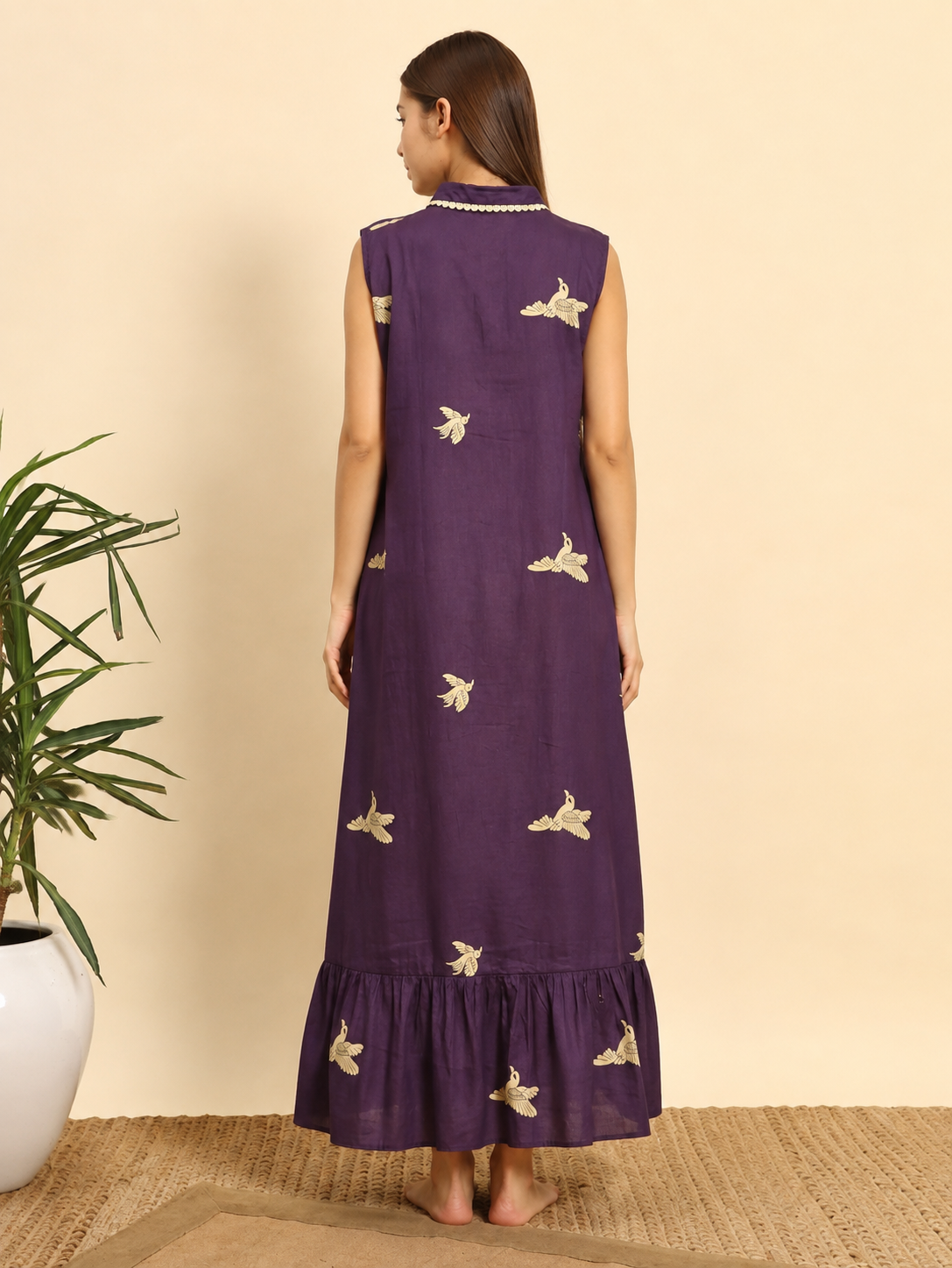 A woman stands with her back to the camera wearing a sleeveless purple maxi dress with gold embroidered birds and a ruffled hem, set against a beige wall with a potted plant on the left.