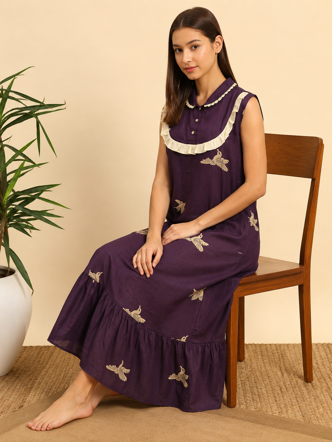 A woman sits on a wooden chair wearing a sleeveless purple maxi dress with cream ruffle trim and embroidered bird motifs. The setting features a beige wall, a woven rug, and a potted plant on the left.