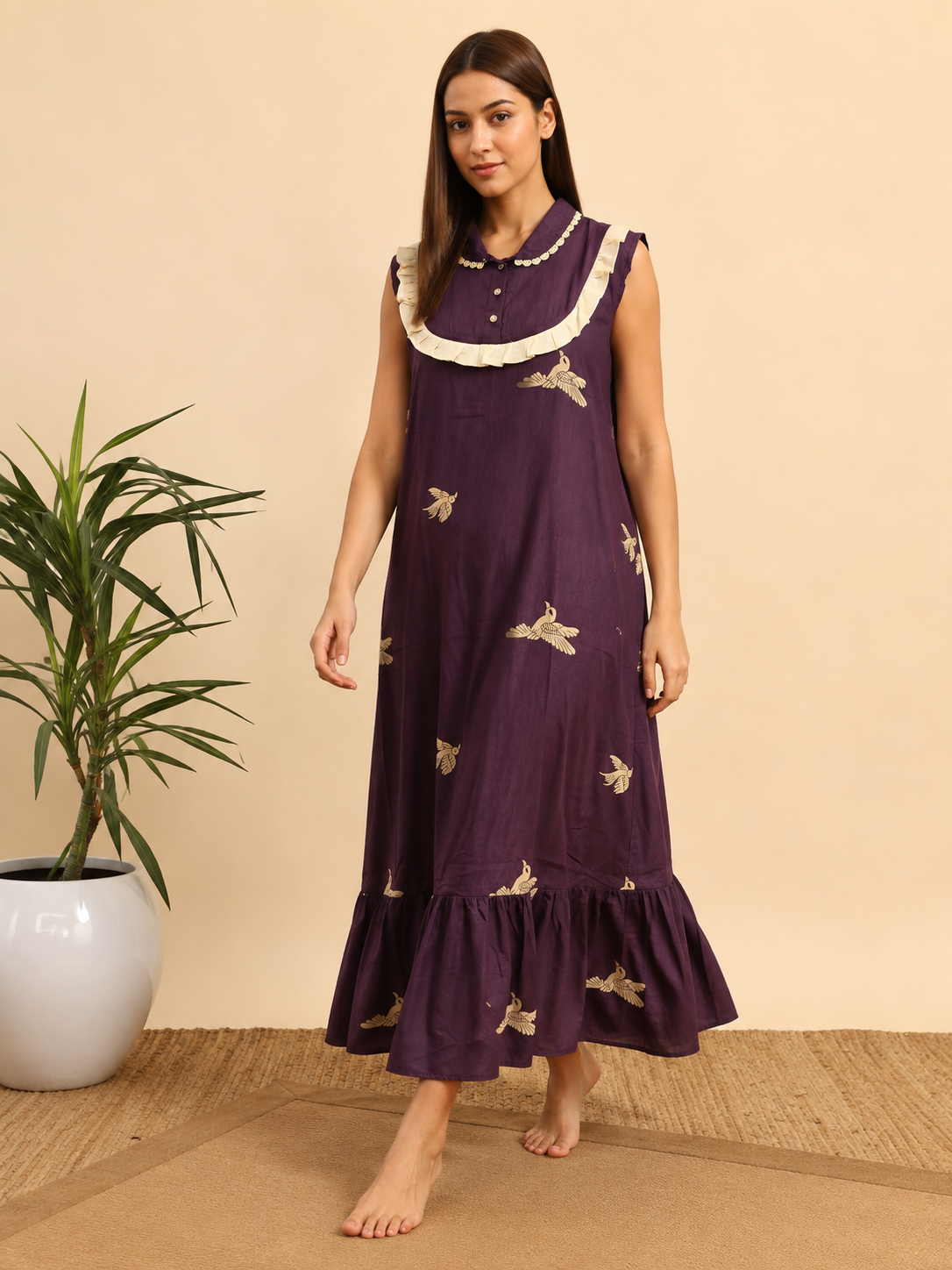 A woman modeling a sleeveless purple maxi dress with cream ruffle trim and gold bird embroidery, standing barefoot in a studio setting with a potted plant on the left.
