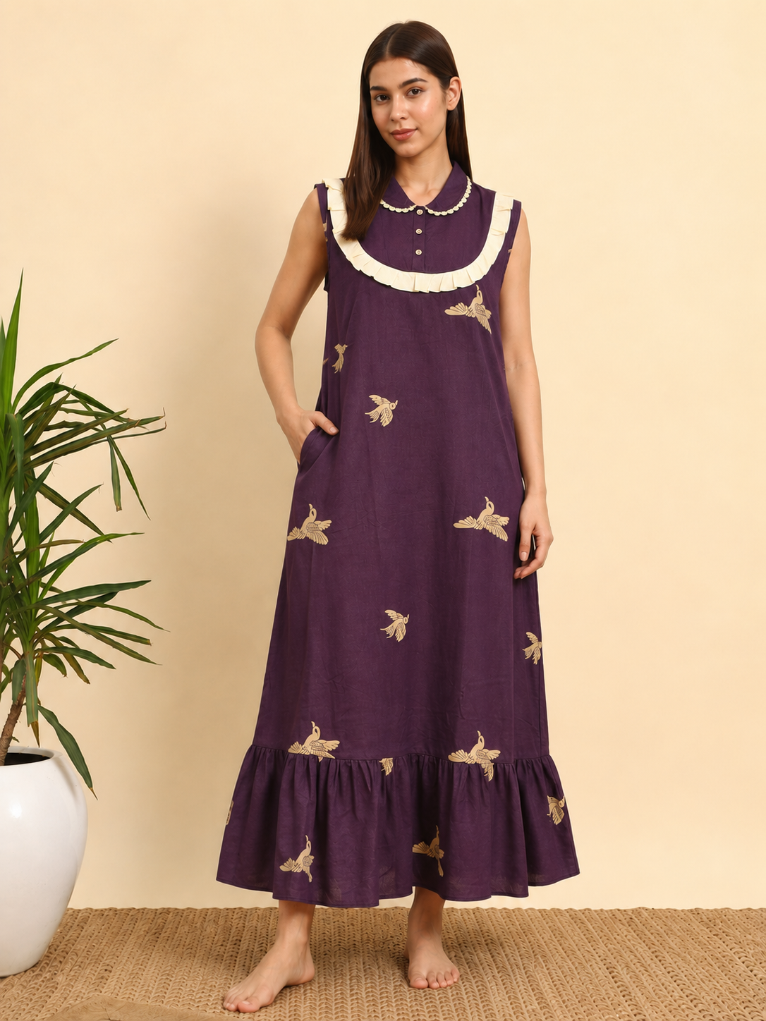 A woman stands barefoot indoors wearing a sleeveless, floor-length purple nightgown with white ruffled trim at the neckline and scattered beige bird motifs. A potted plant and woven rug are visible against a plain beige wall.